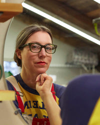 A woman with Bixby Reading Glasses in Heritage Green by CADDIS, featuring a bio-based acetate frame, wearing a yellow t-shirt and blue overalls, stands thoughtfully in a workshop.