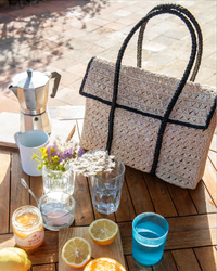 A Violette Flap Bag in Natural/Noir by Maison N.H Paris, on a wooden table outdoors with a stovetop coffee maker, glasses, sliced citrus fruits, and jars of honey nearby.