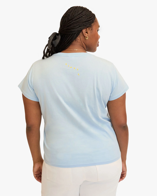 A woman in a Clare V. Oui Classic Tee in Light Blue w/ Cream & Yellow, 100% cotton tee with a subtle yellow graphic on the back, white pants, standing facing away from the camera.