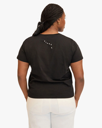 Woman standing with her back to the camera, wearing a black Clare V Block Ciao Classic Tee in Black w/ Cream with a small white text logo and white pants.