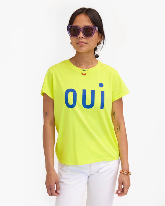 Woman wearing a neon yellow Oui Classic Tee in Neon Yellow w/ Cobalt by Clare V. with the word "oui" in blue letters, paired with white pants and sunglasses.