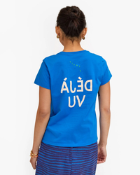A woman standing with her back to the camera, wearing a Clare V. Deja Vu Classic Tee in Bright Cobalt w/ Cream, with the phrase "belau uv" printed in white, reversed letters.