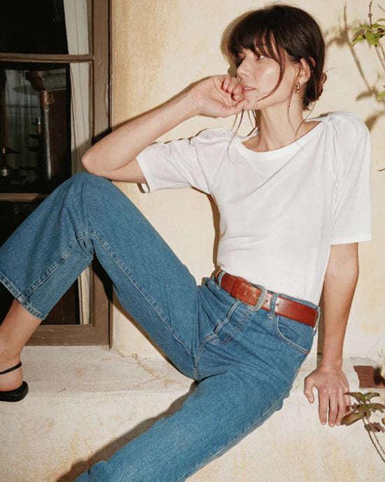 Woman in a casual pose wearing a Nation LTD Deana Solid Envelope T-Shirt in White made of organic cotton and blue jeans with a red belt, sitting by a window.