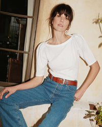 Woman in a white Nation LTD Deana Solid Envelope T Shirt and blue jeans with a red belt, leaning against a wall indoors.
