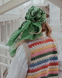 Wearing the Classic Cotton Bandana in Vert by Maison Lecomte Flament, a person with long hair strolls through a white, architectural space, sporting a colorful striped vest made of soft cotton voile.
