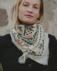 A woman with short blonde hair wears a Maison Lecomte Flament Mallorca Wool & Silk Bandana in creme with green and orange designs around her neck, paired with a dark top, standing against a blurred outdoor background.
