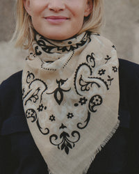 A woman with light hair wears the Classic Wool & Silk Bandana in Beige by Maison Lecomte Flament, featuring a black floral and paisley pattern, draped around her neck. She smiles softly and pairs it with a dark top.