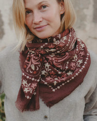 A blonde person wears a light gray button-up sweater and a Maison Lecomte Flament Saint Emilion Wool & Silk Bandana in Rust, standing outdoors against a neutral background.