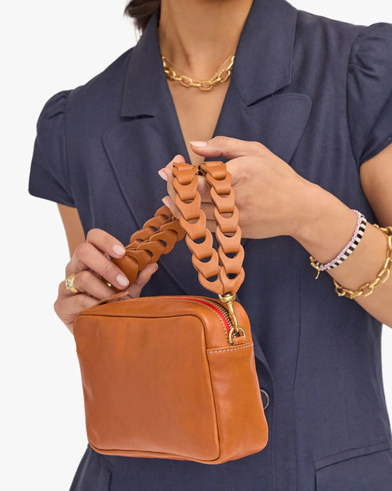 A woman in a blue dress holding a tan Italian leather purse with the Clare V. Double Veg Link Shoulder Strap in Cuoio.