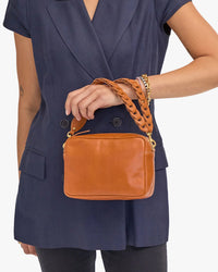 A person in a navy blue dress holding a tan Clare V. Italian leather shoulder bag with a Double Veg Link Shoulder Strap in Cuoio.