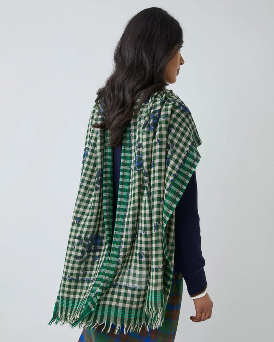 A woman with long, wavy dark hair is seen from the back wearing the Moismont Vichy Wool Scarf No 693 in Flowers British Green, its fringed edges draped over her shoulders, layered atop a navy sweater and checkered skirt.
