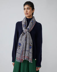 A woman in a navy sweater and green skirt wears a Moismont Vichy Wool Scarf No 693 in Flowers Slate Grey, loosely tied around her neck, standing against a plain background and gazing slightly to the side with a neutral expression.