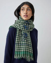 A person wears a navy sweater and the Vichy Wool Scarf No 693 in Flowers British Green by Moismont, featuring fringe, standing against a plain light background and looking slightly to the side.