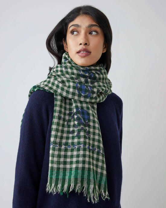 A person wears a navy sweater and the Vichy Wool Scarf No 693 in Flowers British Green by Moismont, featuring fringe, standing against a plain light background and looking slightly to the side.