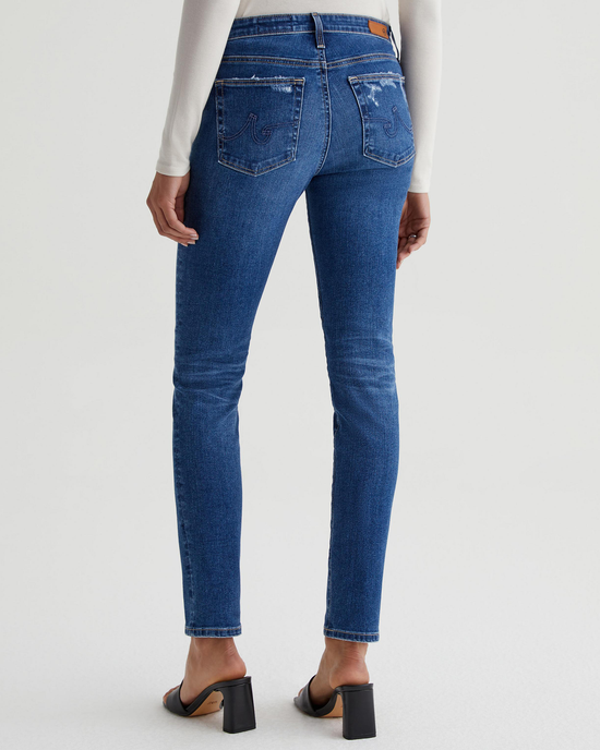 A person wearing blue AG Jeans Prima in Brighton mid-rise jeans and black high-heeled shoes stands with their back to the camera.