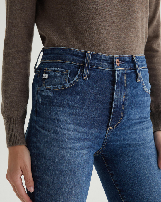 Close-up of a person wearing AG Jeans Farrah Boot in Brighton blue jeans with a brown sweater, hand resting on hip.
