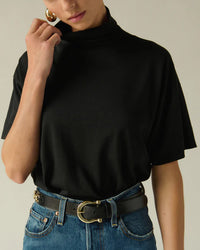 Wearing the Nation LTD Fable Turtleneck Tee in Jet Black, tucked into high-waisted blue jeans with a black belt and gold buckle. Touching the collar and accessorized with large gold hoop earrings.