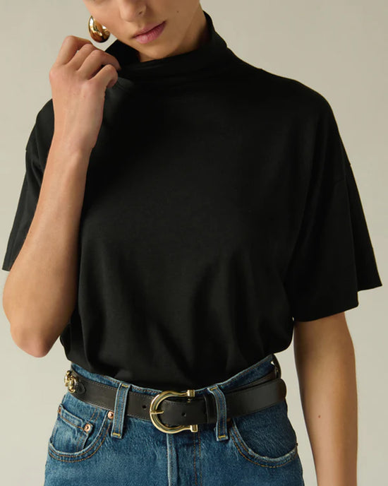 Wearing the Nation LTD Fable Turtleneck Tee in Jet Black, tucked into high-waisted blue jeans with a black belt and gold buckle. Touching the collar and accessorized with large gold hoop earrings.
