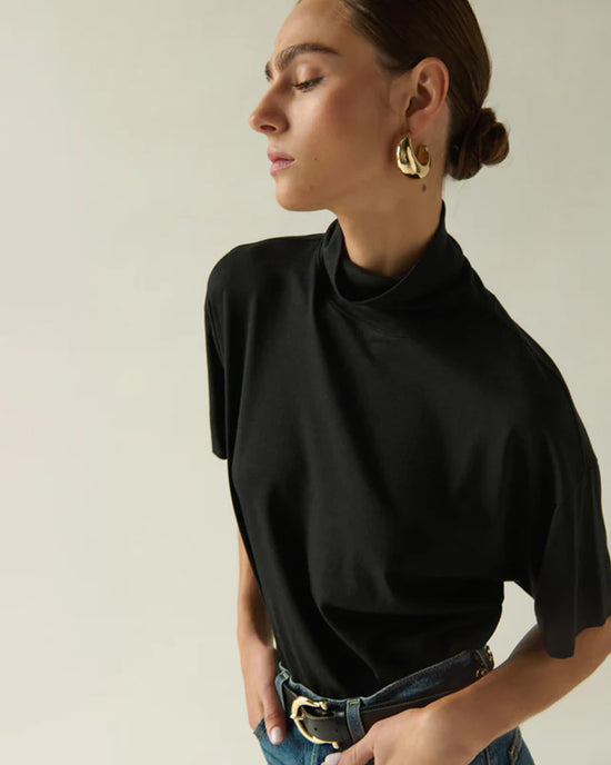 A woman with brown hair in a low bun wears the Nation LTD Fable Turtleneck Tee in Jet Black, gold hoop earrings, and belted blue jeans as she stands against a plain light background, looking to the side with her eyes closed.