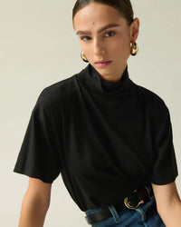A woman with brown hair in a middle-parted style wears large gold hoop earrings, the Nation LTD Fable Turtleneck Tee in Jet Black with a relaxed fit, blue jeans, and a black belt. She poses against a neutral background.