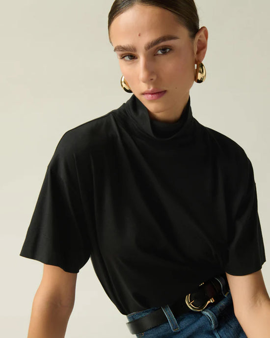 A woman with brown hair in a middle-parted style wears large gold hoop earrings, the Nation LTD Fable Turtleneck Tee in Jet Black with a relaxed fit, blue jeans, and a black belt. She poses against a neutral background.