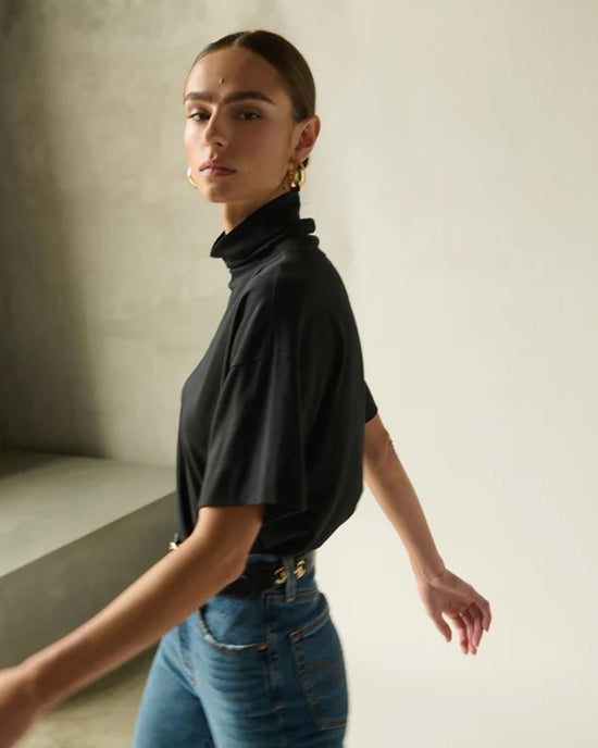 A person with slicked-back hair wears the Nation LTD Fable Turtleneck Tee in Jet Black and blue jeans, standing against a neutral background and looking over their shoulder with one arm extended.