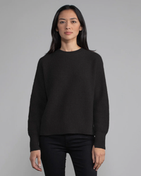 Woman in a black Margaret O'Leary Nora Pullover and black pants standing against a grey background.