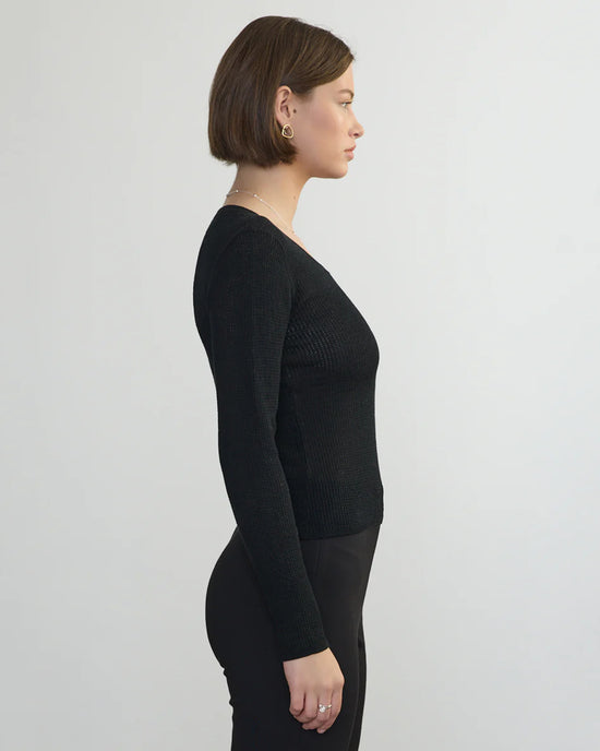 A woman with short brown hair stands in profile against a plain light background, wearing a black Margaret OLeary Scoop Neck Pullover and matching black pants.