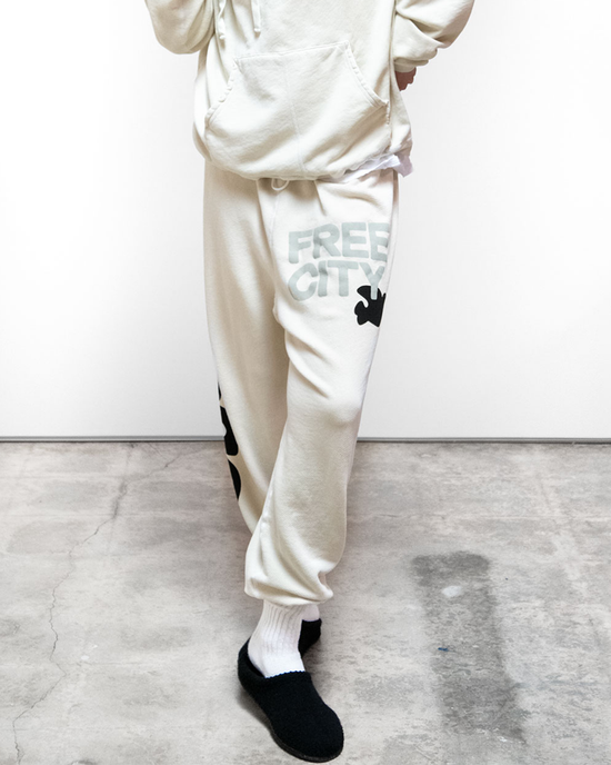 Person standing in cream-colored sweatpants from the medium weight french terry Free City SY OG Freecityletsgo Sweatpant in Bones loungewear set, featuring the text "FREECITY" and a black motif on the left leg, paired with