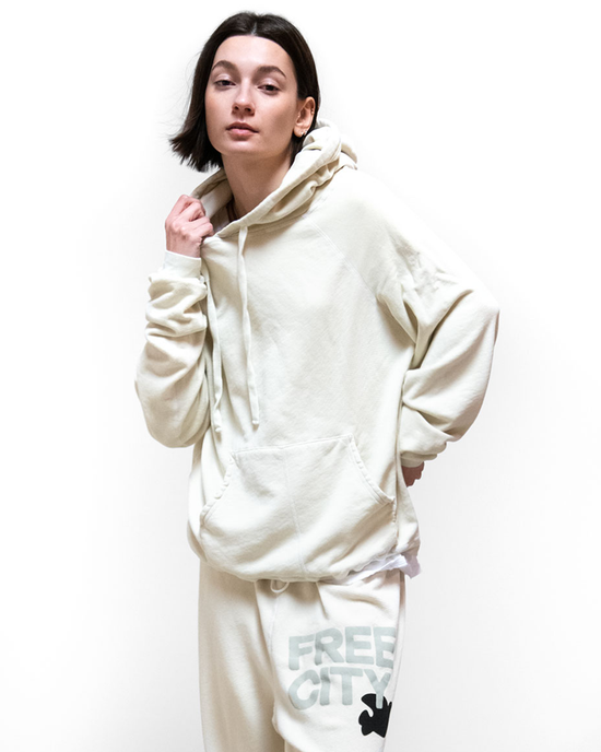 Woman posing in a Free City SY Big Hoodie in Bones loungewear matching set, featuring a beige hoodie and sweatpants made from a promodal/cotton blend against a white background.