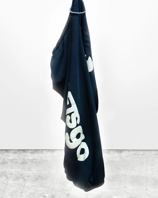 A medium-weight French terry blue hoodie with white text, part of the Free City loungewear matching set, hanging from a hook on a white wall.