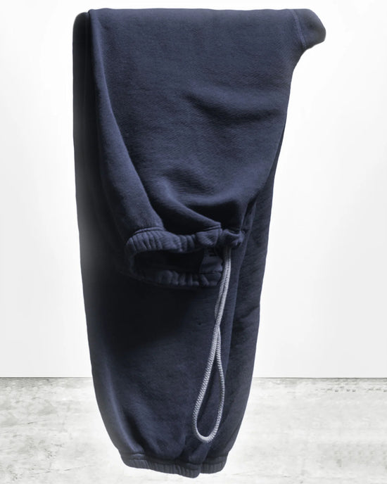 The Free City Basic Goodness OG Sweatpant in Moonshrooms, a pair of navy blue French terry joggers with elastic cuffs and a visible drawstring, is shown folded in half against a plain light gray background.