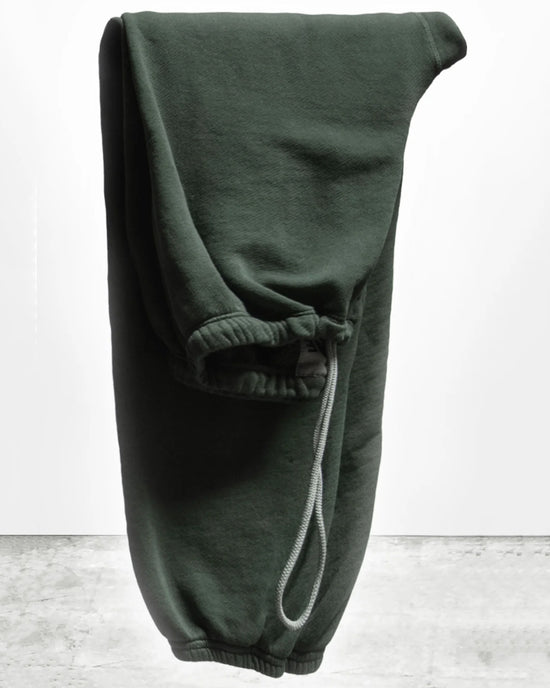 The Free City Basic Goodness OG Sweatpant in Tent, a pair of dark green French terry joggers with elastic cuffs and a drawstring waistband, is shown folded over a hanger against a plain light background.