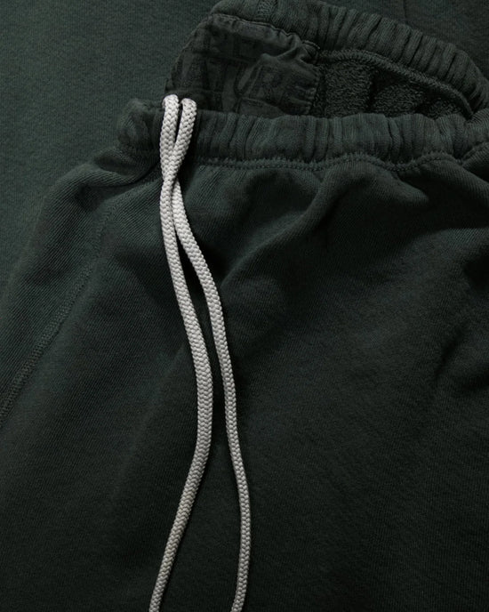 Close-up of Free City Basic Goodness OG Sweatpant in Tent, featuring a dark green French terry fabric, elastic waistband with black text, and white drawstrings laid on top.