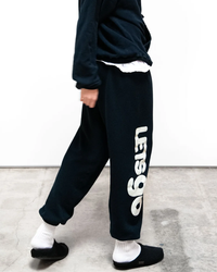 Person wearing black Free City SY OG Freecityletsgo Sweatpant in Deepspace/Cream with a white logo, white socks, and black slippers walking across a concrete floor.