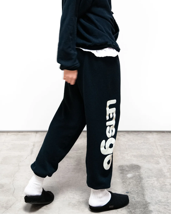 Person wearing black Free City SY OG Freecityletsgo Sweatpant in Deepspace/Cream with a white logo, white socks, and black slippers walking across a concrete floor.