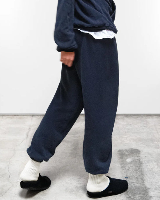 Someone wearing loose dark blue loungewear—a Free City Basic Goodness OG Sweatpant in Moonshrooms with a matching sweatshirt—plus white socks and black slip-ons walks indoors on a concrete floor.