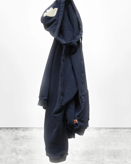 The Free City Basic Goodness Hoodie in Moonshrooms, an oversized navy blue style, hangs from its hood against a white wall. It features a white cloud on the hood and a colorful tag on the pocket for classic brand appeal.