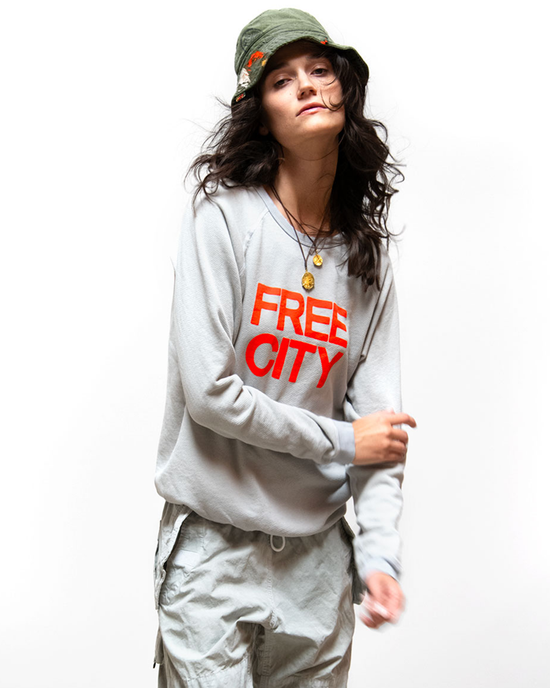 A woman in a Superyumm Biggie Raglan in Dove Grey French Terry sweatshirt with "FREE CITY" in orange letters and a bucket hat, standing against a white background.