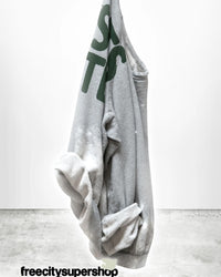 A Free City Artistswanted Raglan in Heather Bleachout, featuring green lettering, hangs against a plain white wall with a concrete floor and "freecitysupershop" in bold black text at the bottom.