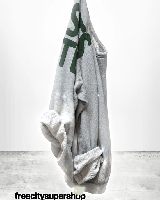 A Free City Artistswanted Raglan in Heather Bleachout, featuring green lettering, hangs against a plain white wall with a concrete floor and "freecitysupershop" in bold black text at the bottom.