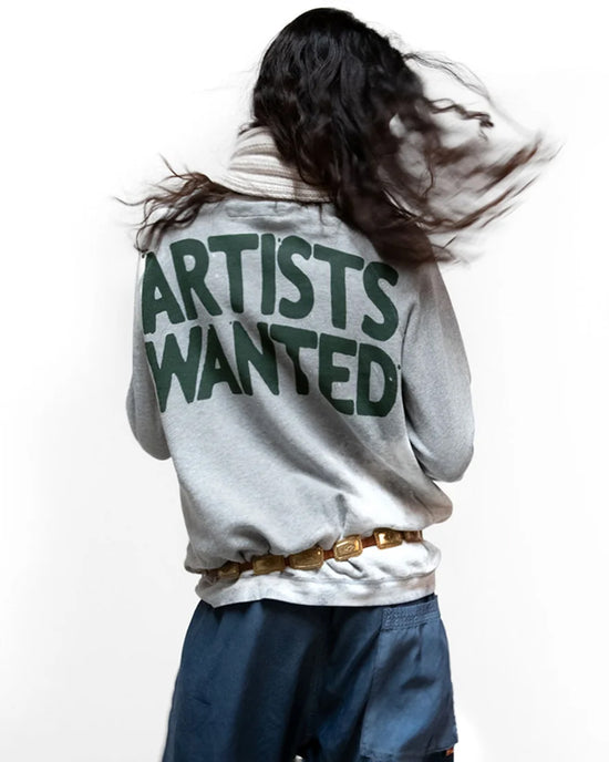 A person with long, dark hair faces away, wearing the Free City Artistswanted Raglan in Heather Bleachout with "ARTISTS WANTED" in large green letters, blue shorts, a gold belt, and white scarf against a plain white background.