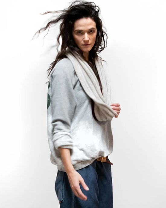 A person with wavy dark hair wears an oversized Artistswanted Raglan in Heather Bleachout by Free City, loose blue pants, and a chunky knit scarf, standing against a plain white background with a windswept look.
