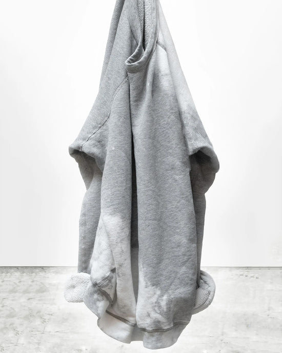 A Free City Rabbit Sweatshirt in Heather Bleachout hangs on a hook against a plain white wall and concrete floor, with the bottom corners slightly curled inward.
