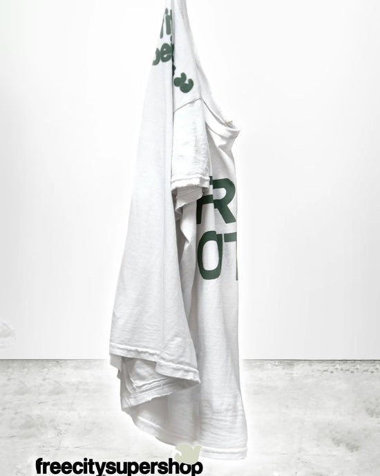 The Free City FC thedoingofitisthebeingofit Supervintage S/S Tee in Laundry Whites, featuring green text, hangs on a hook against a plain white background. "freecitysupershop" appears in black at the bottom of the image.