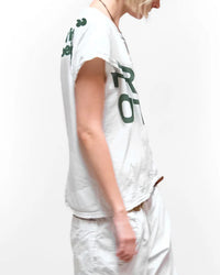 A person with short blonde hair wears the Free City FC thedoingofitisthebeingofit Supervintage S/S Tee in Laundry Whites and white pants, standing against a plain white background, facing slightly to the side with their arm down.