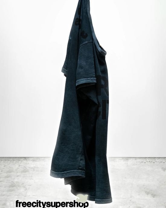 A dark blue FC thedoingofitisthebeingofit Supervintage S/S Tee in Volcano Powder by Free City hangs against a white background. “freecitysupershop” is in black text at the bottom on a gray concrete floor.