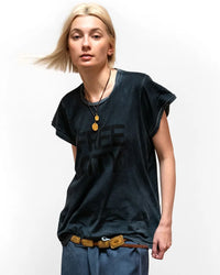 A person with chin-length blonde hair wears a loose, dark Free City FC thedoingofitisthebeingofit Supervintage S/S Tee in Volcano Powder, gold necklaces, blue pants, and a gold-accented belt against a plain white background.