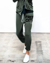 A person standing in a room wearing green Free City Superfluff Lux OG Sweatpants in Bush with "Free City" printed on them and white sneakers, crafted from a promodal/cotton blend.