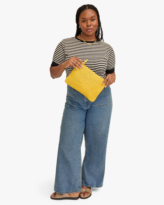Woman in a striped t-shirt and jeans holding a Clare V. Flat Clutch w/ Tabs in Dandelion Rattan, standing against a white background.